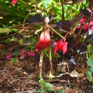 Puede incluir: Una muñeca de hada artesanal con una combinación de colores verde y rojo. La muñeca tiene un tocado, brazos y piernas verdes, con una falda roja en forma de pétalo. El hada sostiene una flor roja en un jardín.
