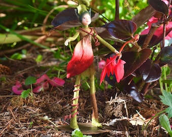 May include: A handcrafted fairy doll with a green and red color scheme. The doll has a green headpiece, arms, and legs, with a red petal-shaped skirt. The fairy is holding a red flower, standing in a garden setting.
