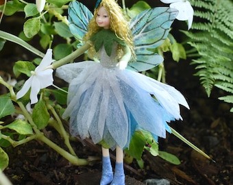 May include: A miniature fairy figurine with blonde hair, blue wings, and a blue and white dress. The fairy is surrounded by green foliage and white flowers. The text "Fae Folk World.com" is at the bottom.