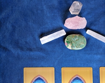 Op de afbeelding: Twee gouden tarotkaarten met blauwe en witte ontwerpen op een blauwe achtergrond. De kaarten hebben een blauw en wit ontwerp met een symbool van een cirkel met vleugels erin. De kaarten zijn omringd door kristallen, waaronder een helder kristal, een roze kristal en een groen kristal.