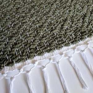 May include: A green woven rug with a white fringe. The rug has a textured surface and is made of natural fibers.