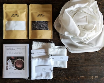 Op de afbeelding: Een natuurverfset met diverse componenten. De set bevat twee kleine bruine papieren zakken met de merknaam "ANINI", een boekje met de tekst "NATURAL DYE KIT", stofstalen en een witte sjaal. De stofstalen zijn gelabeld met "Bamboo-Rayon", "Cotton" en "Silk".