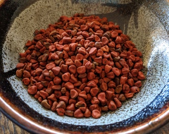 May include: A small bowl filled with red annatto seeds. The bowl is brown with a white speckled glaze.