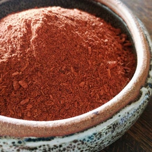 May include: A close-up of a small ceramic bowl filled with a reddish-brown powder. The bowl has a speckled glaze and a brown rim.