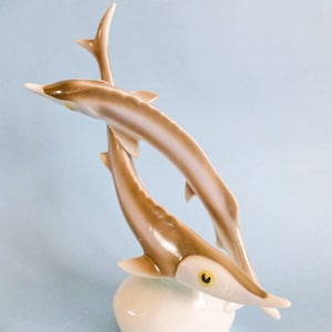 Könnte beinhalten: Eine Keramik-Delfinfigur in Braun- und Weißtönen. Die Skulptur zeigt zwei nach oben springende Delfine, die an der Basis verbunden sind. Die Delfine haben detaillierte Flossen und Augen, und die Basis ist eine abgerundete, cremefarbene Form.