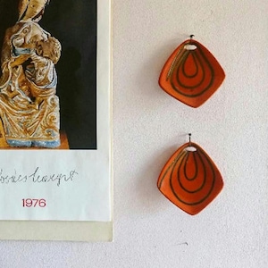 Può includere: Un calendario da parete con l'immagine di una scultura religiosa con il testo 1976 e due piatti da parete in ceramica arancione a forma di diamante con motivi a goccia concentrici verde scuro appesi verticalmente.
