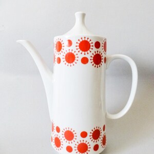May include: White ceramic coffee pot with a tall, slender design. The pot features a red sunburst pattern around the top and bottom. It has a curved spout, a handle, and a lid with a small knob.