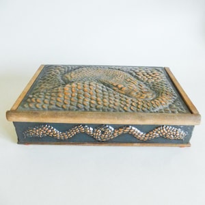 May include: A rectangular box with a detailed embossed design. The lid features a central design with a textured, scale-like pattern in shades of orange and green. The box has a dark blue base with a decorative border.
