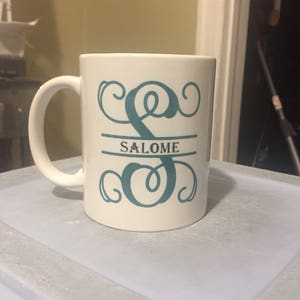 May include: A white ceramic mug featuring a large, teal-colored decorative letter S with ornate swirls. A horizontal line cuts through the center of the letter, containing the name SALOME printed in a dark, capitalized serif font.