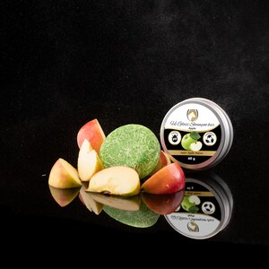 Op de afbeelding: Een groene vaste shampoo-bar met een label met de tekst "Old Spice Shampoo Bar Apple" en een groene appelafbeelding. De bar is omringd door appelschijfjes op een zwart reflecterend oppervlak.