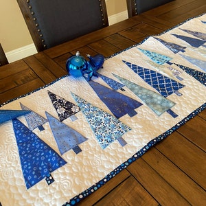 May include: A blue and white quilted table runner with a row of blue Christmas trees. The runner is on a wooden table with blue ornaments.