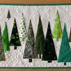 May include: A quilted Christmas table runner featuring a forest of green trees in various patterns and shades. The trees are set against a white background with a swirling pattern and a green border. A festive home decor item.