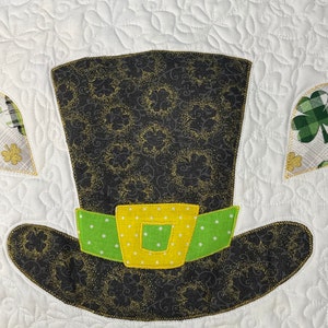 May include: A black and green felt leprechaun hat with a gold buckle and white polka dots on the band. The hat is decorated with a gold glitter pattern.