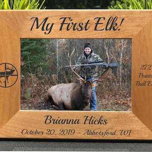 May include: A wooden photo frame with a personalized inscription that reads "My First Elk!" The frame features a photo of a woman with a rifle and a large elk. The inscription also includes the hunter's name, the date of the hunt, and the location. The text "272 Points Bull Elk" is also included.
