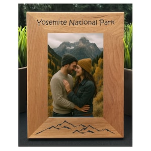 May include: Wooden photo frame with the text "Yosemite National Park" at the top. The frame holds a photo of a couple in front of a mountain landscape. The bottom of the frame features a mountain range design.