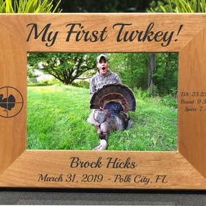 May include: A wooden picture frame with a photo of a young person holding a wild turkey. The frame is engraved with the text "My First Turkey!" and the hunter's name, date, and location. The turkey's weight, beard length, and spur length are also engraved on the frame.