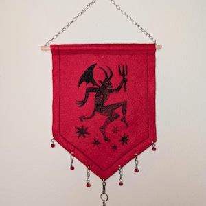 May include: A red felt wall banner featuring a black screen-printed illustration of a horned devil figure holding a pitchfork, surrounded by small star shapes. The banner hangs from a wooden dowel with a silver chain and has red bead accents.