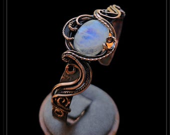 May include: A handcrafted wire-wrapped ring featuring a large, oval, iridescent moonstone. The ring is made with copper-colored wire and has intricate detailing around the stone. The ring is displayed on a gray velvet ring holder.