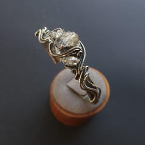Pu&ograve; includere: Un bracciale a polsino argentato con un grande cristallo trasparente di forma irregolare e due cristalli pi&ugrave; piccoli. I cristalli sono avvolti in modo intricato con filo metallico, creando un design artistico unico. Il bracciale &egrave; esposto su un supporto di legno su sfondo scuro.