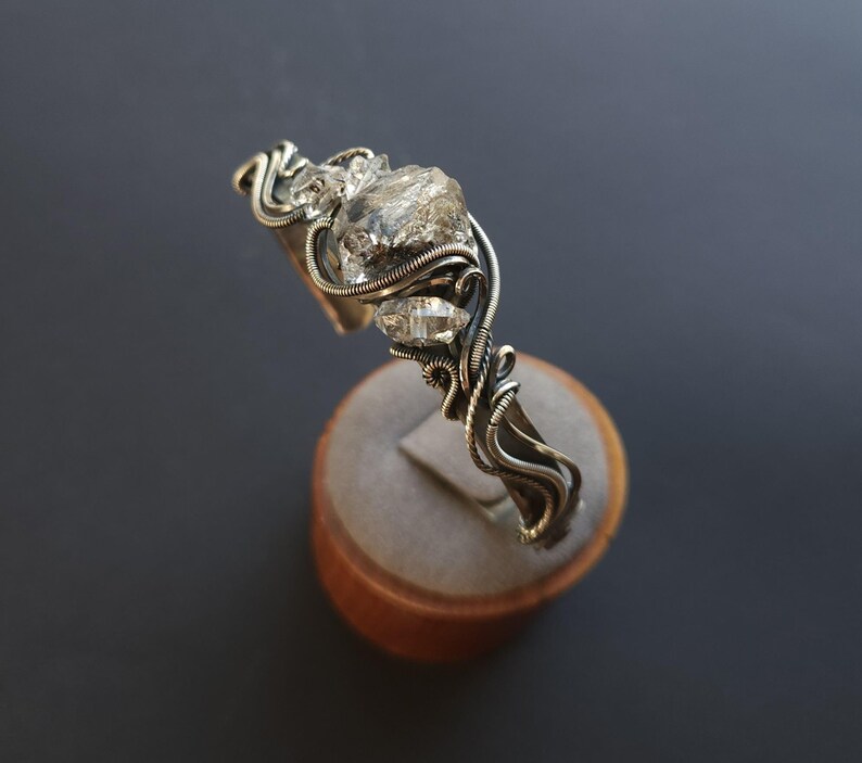 Pu&ograve; includere: Un bracciale a polsino argentato con un grande cristallo trasparente di forma irregolare e due cristalli pi&ugrave; piccoli. I cristalli sono avvolti in modo intricato con filo metallico, creando un design artistico unico. Il bracciale &egrave; esposto su un supporto di legno su sfondo scuro.