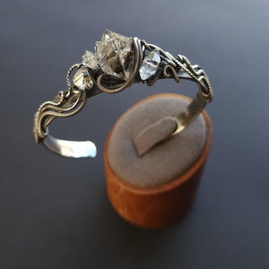 Pu&ograve; includere: Un bracciale a polsino in argento con un gruppo di cristalli di quarzo trasparente. Il bracciale presenta un design dettagliato con filo metallico e una finitura argentata lucida. I cristalli sono il punto focale, aggiungendo un tocco naturale ed elegante al gioiello.