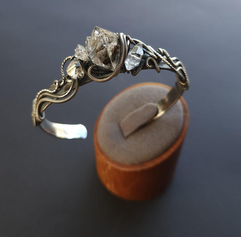 Pu&ograve; includere: Un bracciale a polsino in argento con un gruppo di cristalli di quarzo trasparente. Il bracciale presenta un design dettagliato con filo metallico e una finitura argentata lucida. I cristalli sono il punto focale, aggiungendo un tocco naturale ed elegante al gioiello.