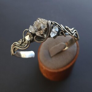 Pu&ograve; includere: Un bracciale a polsino argentato con cristalli di quarzo trasparente. I cristalli sono incastonati in un design avvolto in filo metallico, con una fascia in argento lucido. Il bracciale &egrave; esposto su un supporto marrone su sfondo scuro.