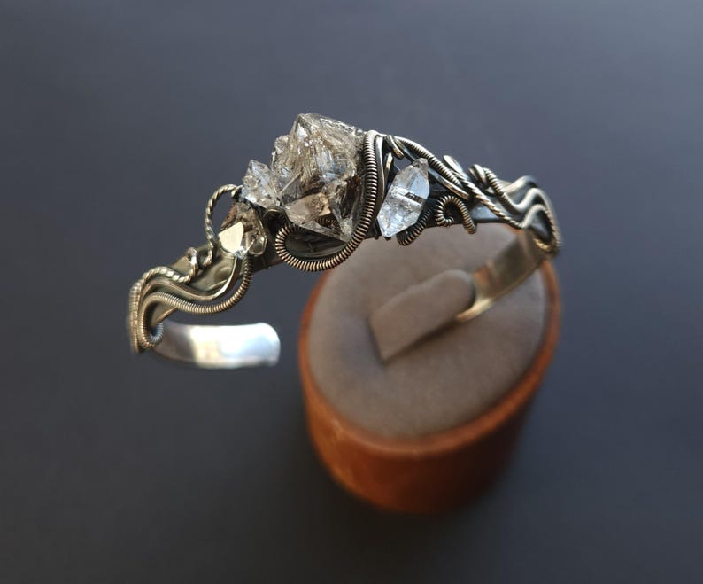 Pu&ograve; includere: Un bracciale a polsino argentato con cristalli di quarzo trasparente. I cristalli sono incastonati in un design avvolto in filo metallico, con una fascia in argento lucido. Il bracciale &egrave; esposto su un supporto marrone su sfondo scuro.