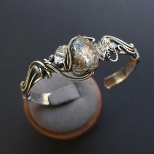 Pu&ograve; includere: Un bracciale a polsino in argento fatto a mano con tre cristalli di quarzo grezzi e trasparenti. I cristalli sono avvolti in filo metallico con un design intricato. Il bracciale &egrave; esposto su un espositore di gioielli in velluto grigio.
