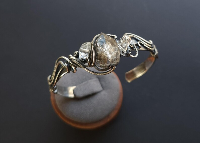 Pu&ograve; includere: Un bracciale a polsino in argento fatto a mano con tre cristalli di quarzo grezzi e trasparenti. I cristalli sono avvolti in filo metallico con un design intricato. Il bracciale &egrave; esposto su un espositore di gioielli in velluto grigio.