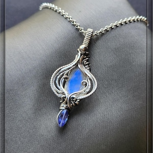 May include: A silver pendant necklace with a teardrop-shaped, iridescent blue gemstone. The pendant is intricately wire-wrapped with a delicate design, and the necklace has a silver chain. The text "Wire Jewelry by Shawna. Etsy.com" is at the bottom.