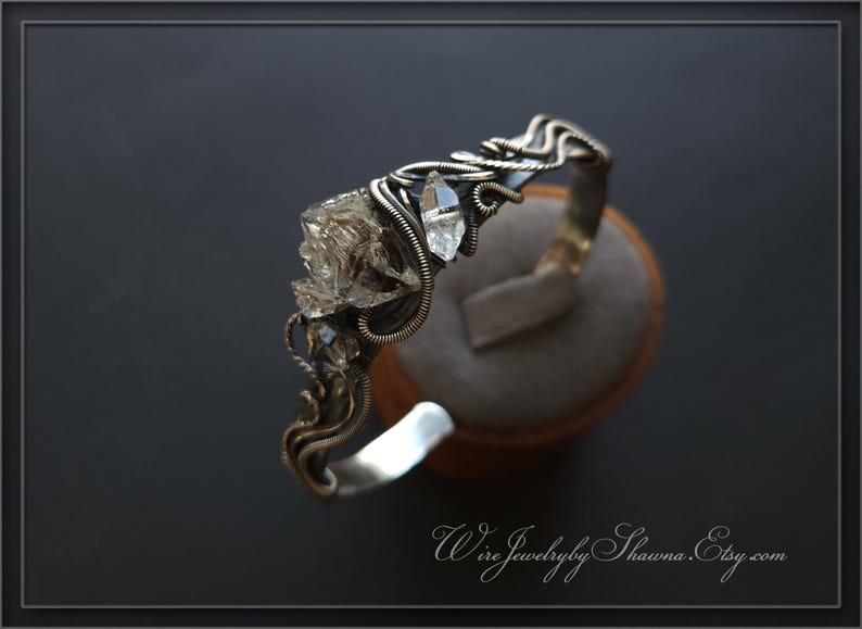 Pu&ograve; includere: Un bracciale a polsino argentato fatto a mano con un'intricata filigrana e accenti di cristallo trasparente. Il bracciale presenta un gruppo di cristalli e un singolo cristallo appuntito. Il design &egrave; organico ed elegante.