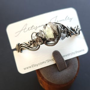 Pu&ograve; includere: Un braccialetto artigianale in filo argentato con accenti di cristalli grezzi. Il braccialetto &egrave; esposto su una carta bianca con il testo "Artisan Jewelry" e "jewelrybyShawna". &Egrave; visibile anche l'URL del negozio Etsy.