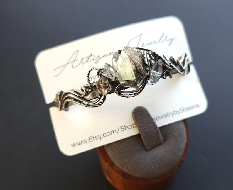 Pu&ograve; includere: Un braccialetto artigianale in filo argentato con accenti di cristalli grezzi. Il braccialetto &egrave; esposto su una carta bianca con il testo "Artisan Jewelry" e "jewelrybyShawna". &Egrave; visibile anche l'URL del negozio Etsy.