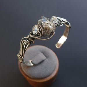 Pu&ograve; includere: Un bracciale a polsino argentato con tre cristalli di quarzo trasparente. I cristalli sono avvolti in filo metallico e fissati su una fascia curva. Il bracciale &egrave; esposto su un espositore per gioielli in velluto grigio.