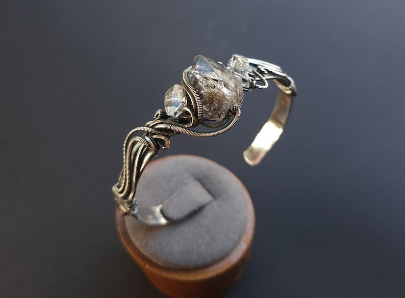 Pu&ograve; includere: Un bracciale a polsino argentato con tre cristalli di quarzo trasparente. I cristalli sono avvolti in filo metallico e fissati su una fascia curva. Il bracciale &egrave; esposto su un espositore per gioielli in velluto grigio.