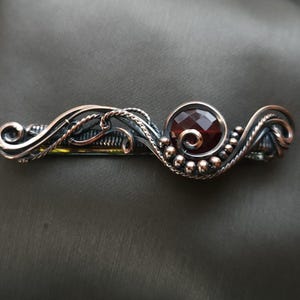 Può includere: Fermaglio per capelli fatto a mano con una gemma centrale rotonda, sfaccettata, rosso scuro. La lavorazione del metallo presenta un'intricata filigrana in argento e rame, con piccole perle d'argento che accentuano il design. Il fermaglio è progettato per fissare i capelli.