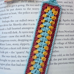 May include: A crocheted bookmark with a blue, yellow, and red pattern. The bookmark is made with a granny square stitch and has a fringe on the top.