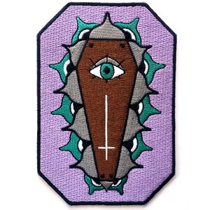 May include: An embroidered patch with a purple background featuring a brown coffin with a white cross and a green eye. The coffin is surrounded by a ring of gray and green thorns.