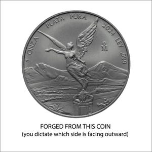 The image is a silver coin with an embossed figure of a winged woman, standing on a pedestal with mountains in the background. The coin is described as "FORGED FROM THIS COIN" and "you dictate which side is facing outward".