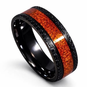 Puede incluir: Un anillo negro con una banda central de veta de madera marrón rojizo y bordes negros decorativos. El anillo tiene un acabado pulido. La veta de la madera es marrón rojizo.