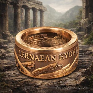 A gold-colored ring with an embossed design of a snake and the text "Lernaean Hydra" on it, set against a rocky landscape with ancient ruins in the background.