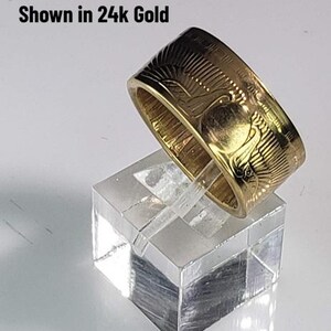 May include: A gold ring with an engraved design, shown in 24k gold. The ring is displayed on a clear, square stand. The ring's design features a bird and sunburst pattern. The ring is a band style.