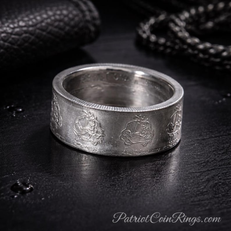 A silver-colored ring with embossed designs on a black surface, with a black leather item and a chain visible in the background.