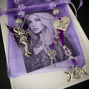 May include: A purple organza drawstring bag containing a card with a black and white illustration of a woman. The bag is adorned with a beaded strand featuring iridescent beads, a silver fairy charm, a heart charm with the text Blessed Be, a key charm, and a triple moon pentacle charm.