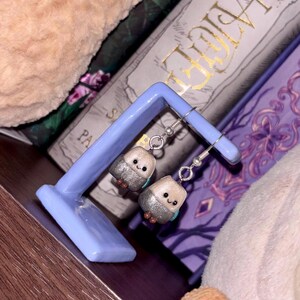 May include: A pair of handmade bird-shaped earrings with silver glitter accents and teal details, hanging from silver hooks. The earrings are displayed on a light blue stand, with books and plush toys in the background.