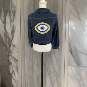 May include: A blue denim jacket with a cropped length and a large embroidered evil eye design on the back.