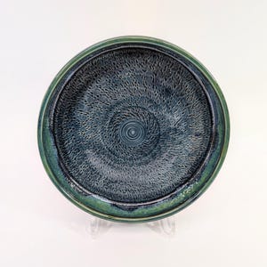 Pode incluir: Um prato redondo de cerâmica com um esmalte texturizado azul escuro e uma borda verde. O prato tem um padrão de espiral no centro.