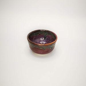 May include: A small, round ceramic bowl with a dark purple and green glaze. The bowl has a slightly uneven rim and a smooth, glossy finish.