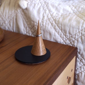 May include: A wooden ring holder with a black base and a pointed cone. A silver ring is placed on the cone.